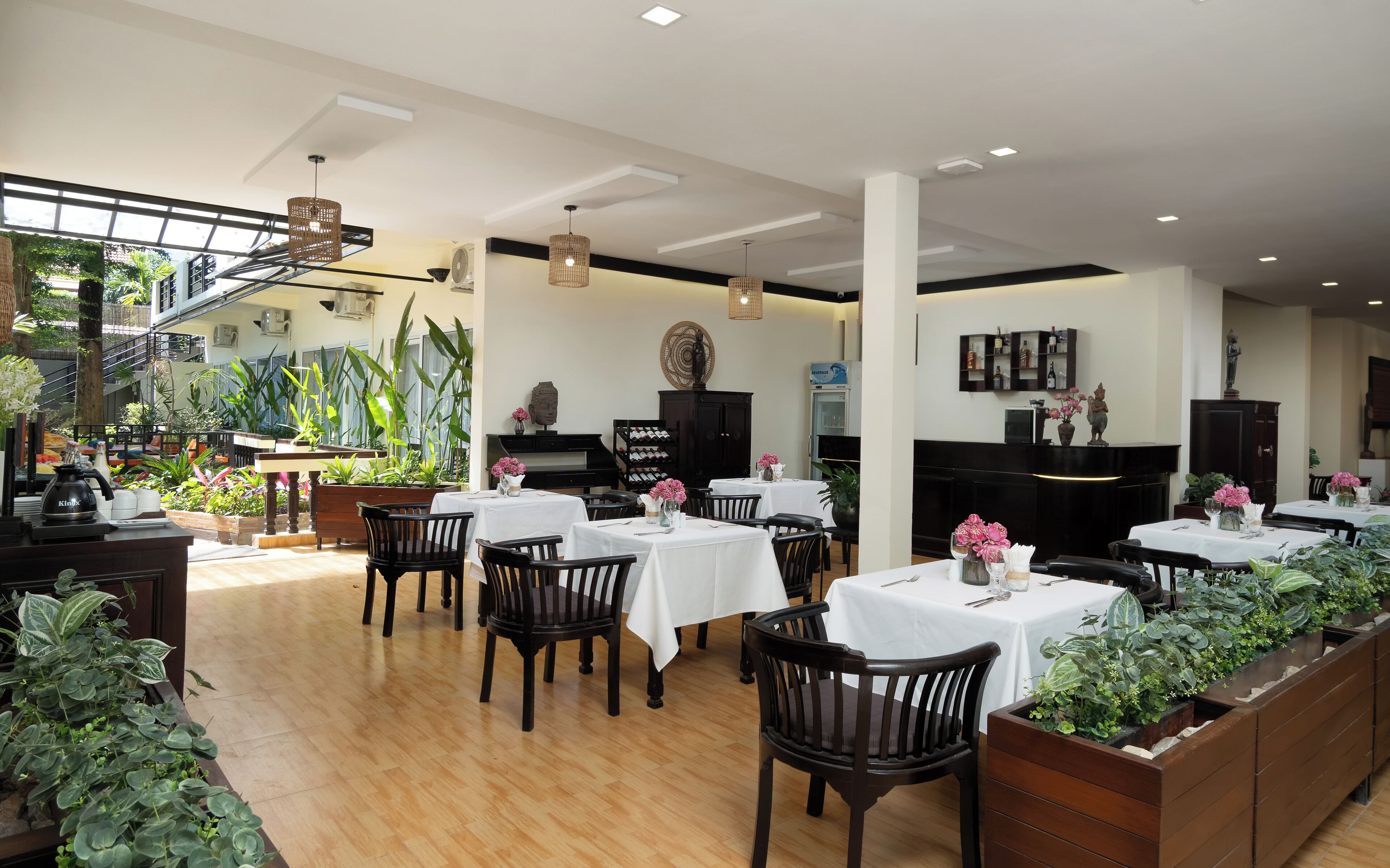 A restaurant with multiple tables and chairs, indoor plants, and a staircase leading to an upper level.