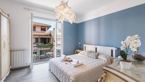 A bedroom with a bed, a nightstand, a dresser, a hanging light fixture, a window with a view of buildings, and a balcony with chairs and plants. - Incanto Del Lago 50m to Lake Maggiore, Baveno, Italy (Baveno)