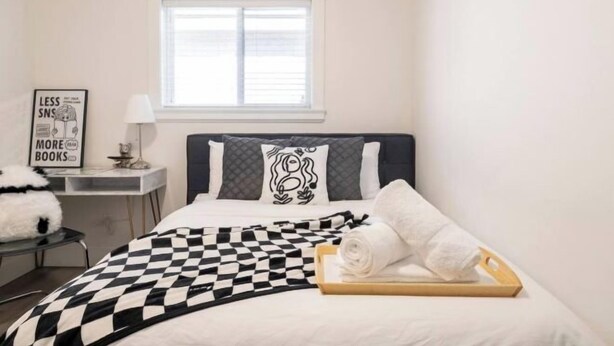 A bedroom with a checkered bedspread, a bedside table with a lamp, a framed quote on the wall, and a window above the bed. - Sweet Home-Separate Entry-Near YVR Airport & Skytrain & Costco (Greater Vancouver)