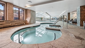 An indoor swimming pool with clear glass walls, stone flooring, and a seating area with benches.