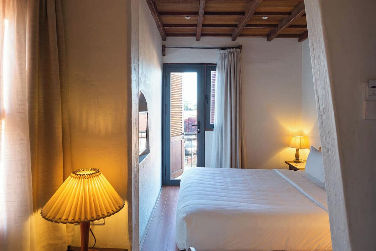 Une chambre d’hôtel chaleureuse avec un plafond en bois, un lit recouvert de draps blancs, une table de chevet avec une lampe et une vue sur l’extérieur par une porte ouverte.
