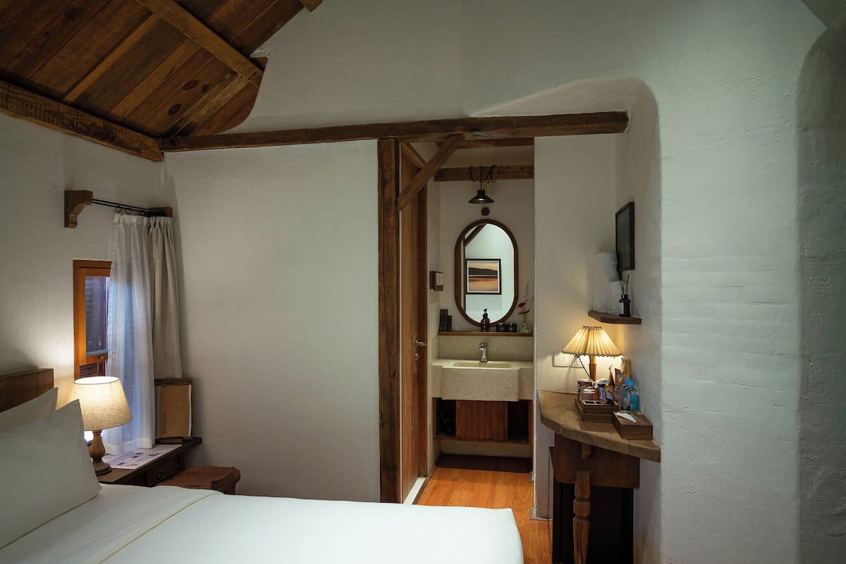 Une chambre avec un plafond en bois, un lit avec une literie blanche, une table de chevet avec une lampe, et une salle de bain avec un meuble-lavabo en bois, un lavabo et un miroir.
