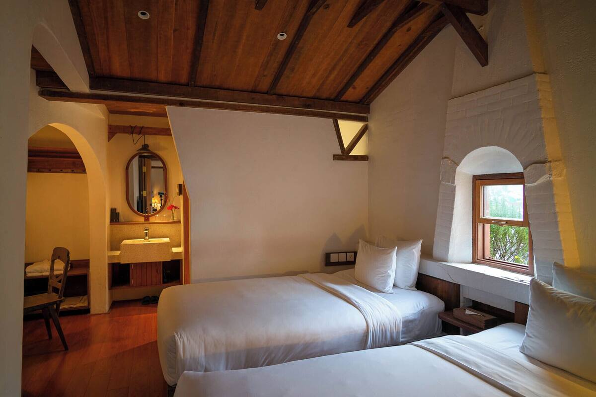 Une chambre avec deux lits, un plafond en bois, une fenêtre et une salle de bain avec un lavabo et un miroir.