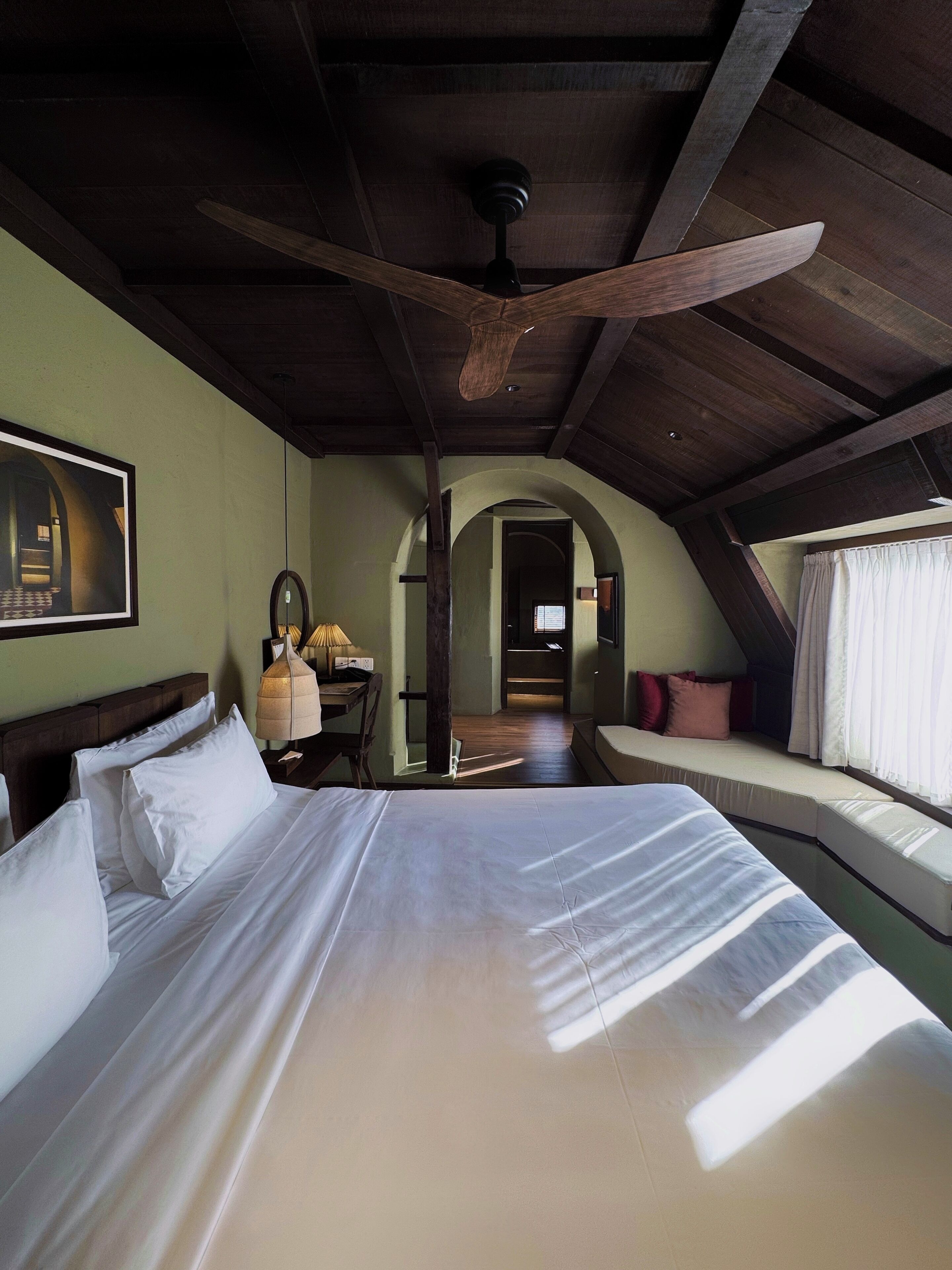 A bedroom with a large bed, a desk with a lamp, and a view of a hallway.