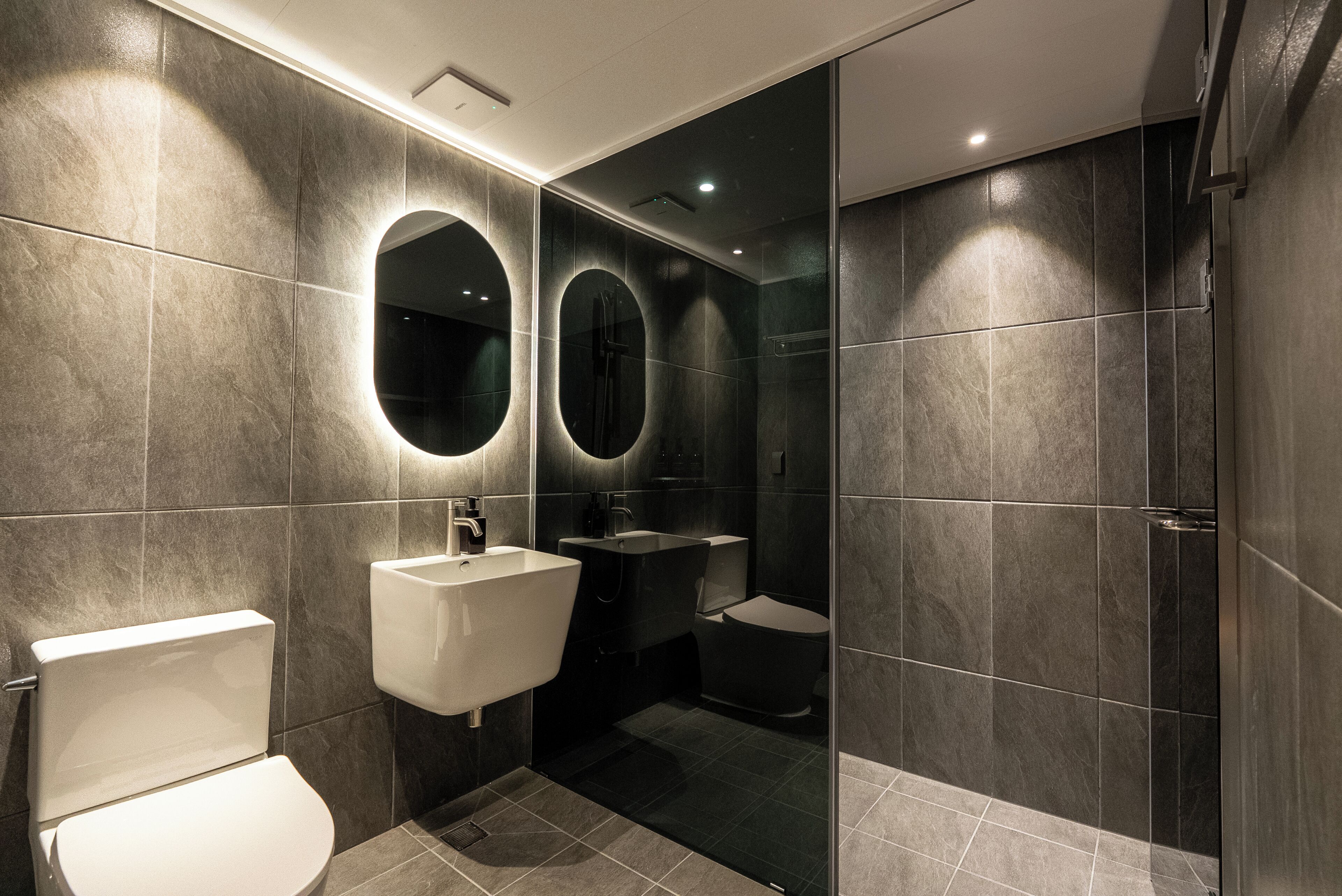 A modern bathroom with a toilet, sink, and shower area, featuring dark tiles and a glass partition.