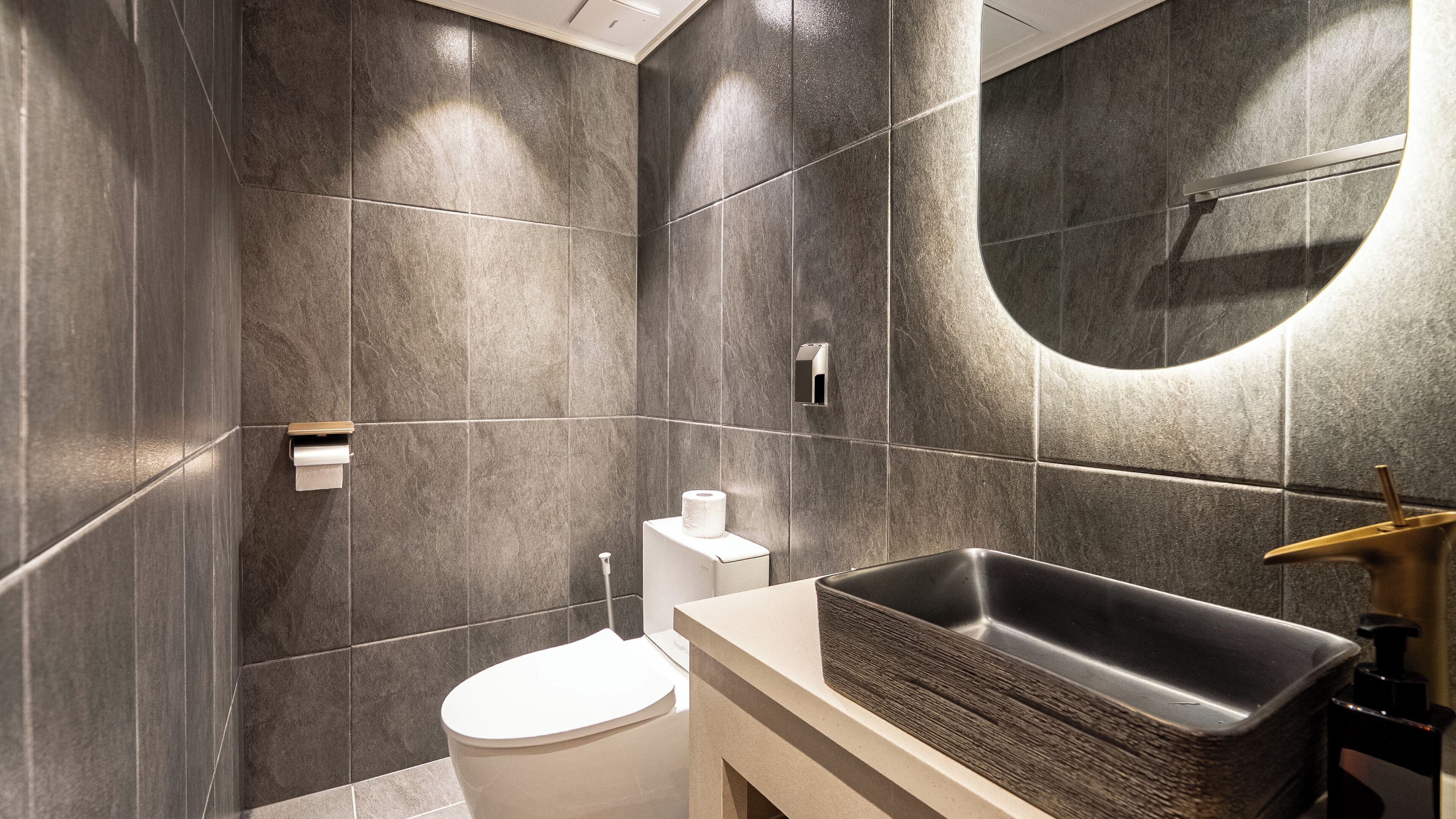 Ein modernes Badezimmer mit dunklen Fliesen, einer weißen Toilette, einem Waschbecken mit Holztablett und einem Spiegel mit rundem Rahmen.