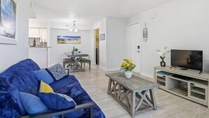 Een woonkamer met een blauwe bank, een houten salontafel, een tv-meubel en een eethoek op de achtergrond.