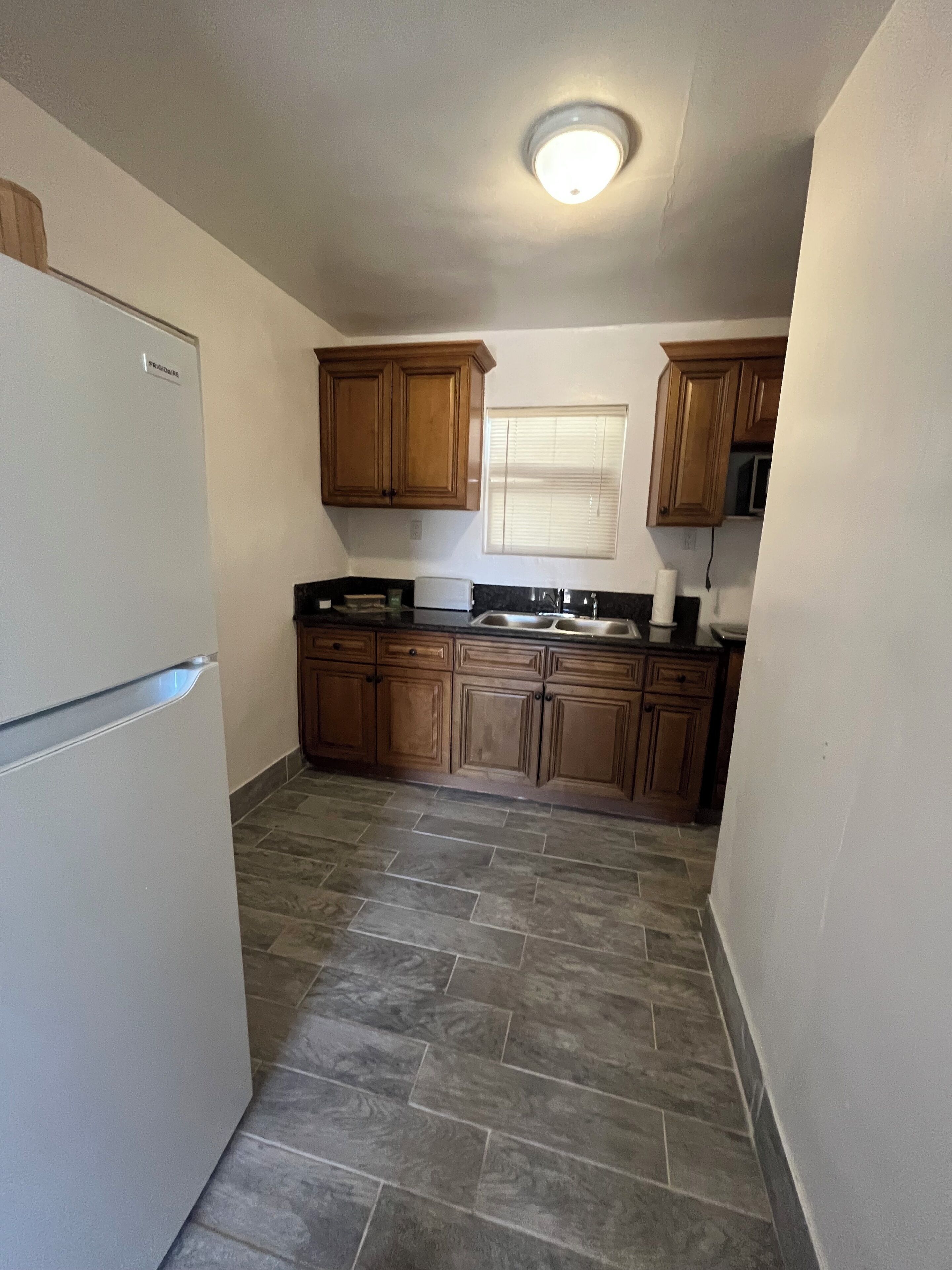 A compact kitchen with wooden cabinets, a refrigerator, a window with blinds, and a tiled floor.