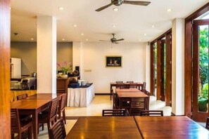 A dining area with wooden tables and chairs, a ceiling fan, and a view of greenery through large windows. - Sabaidee@Lao Hotel Vientiane (Vientiane)