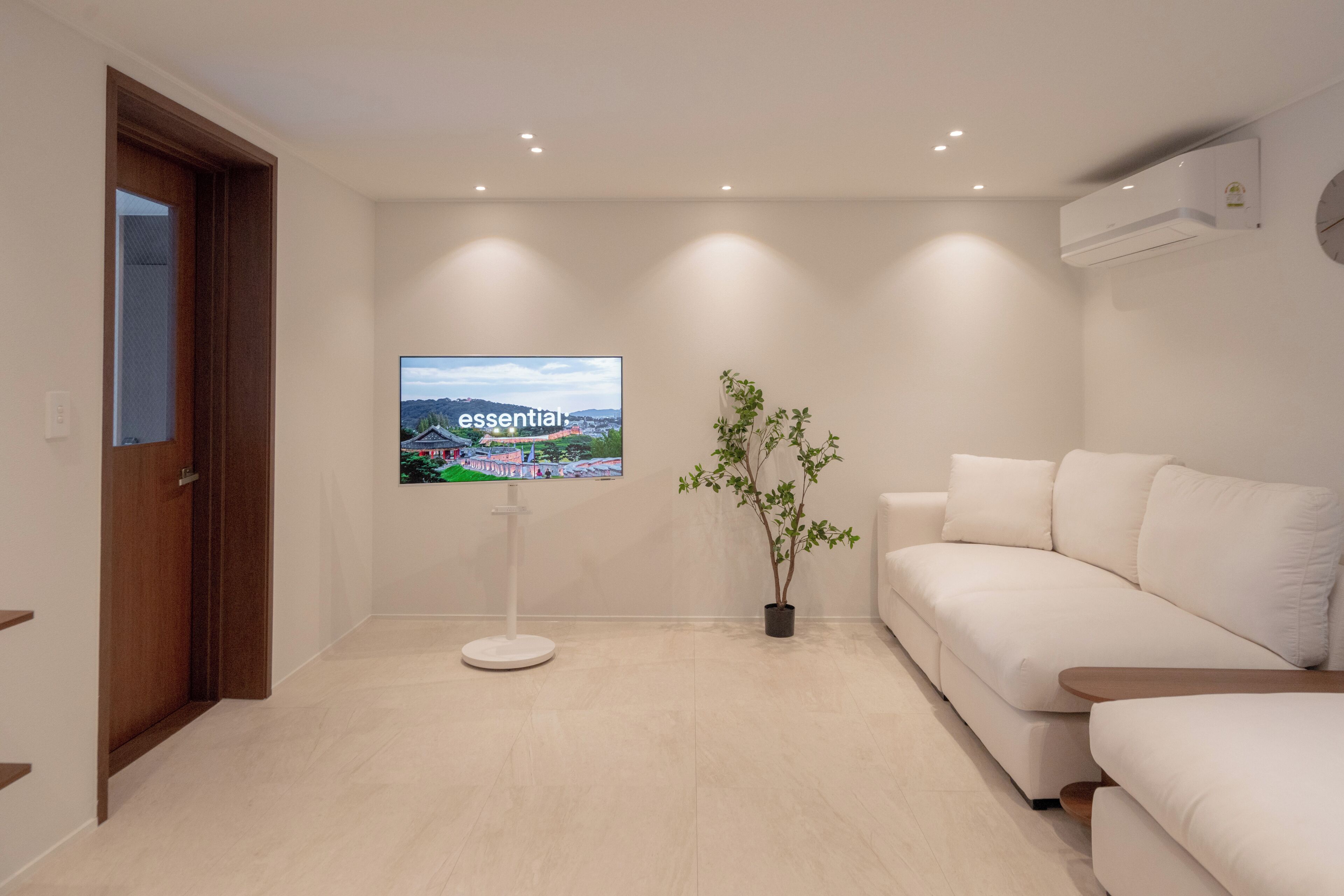 A modern living room with a white sofa, a flat-screen TV mounted on the wall, a potted plant, and recessed ceiling lighting.