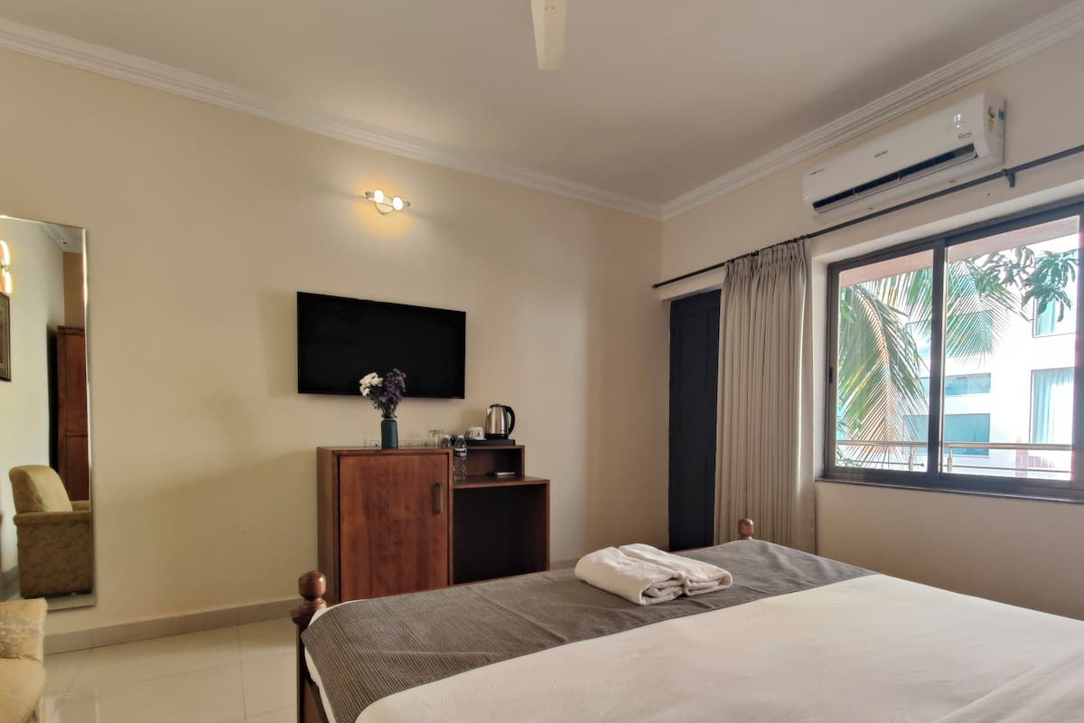 A hotel room with a bed, a wooden cabinet, a flat-screen TV, a window with curtains, and an air conditioning unit.