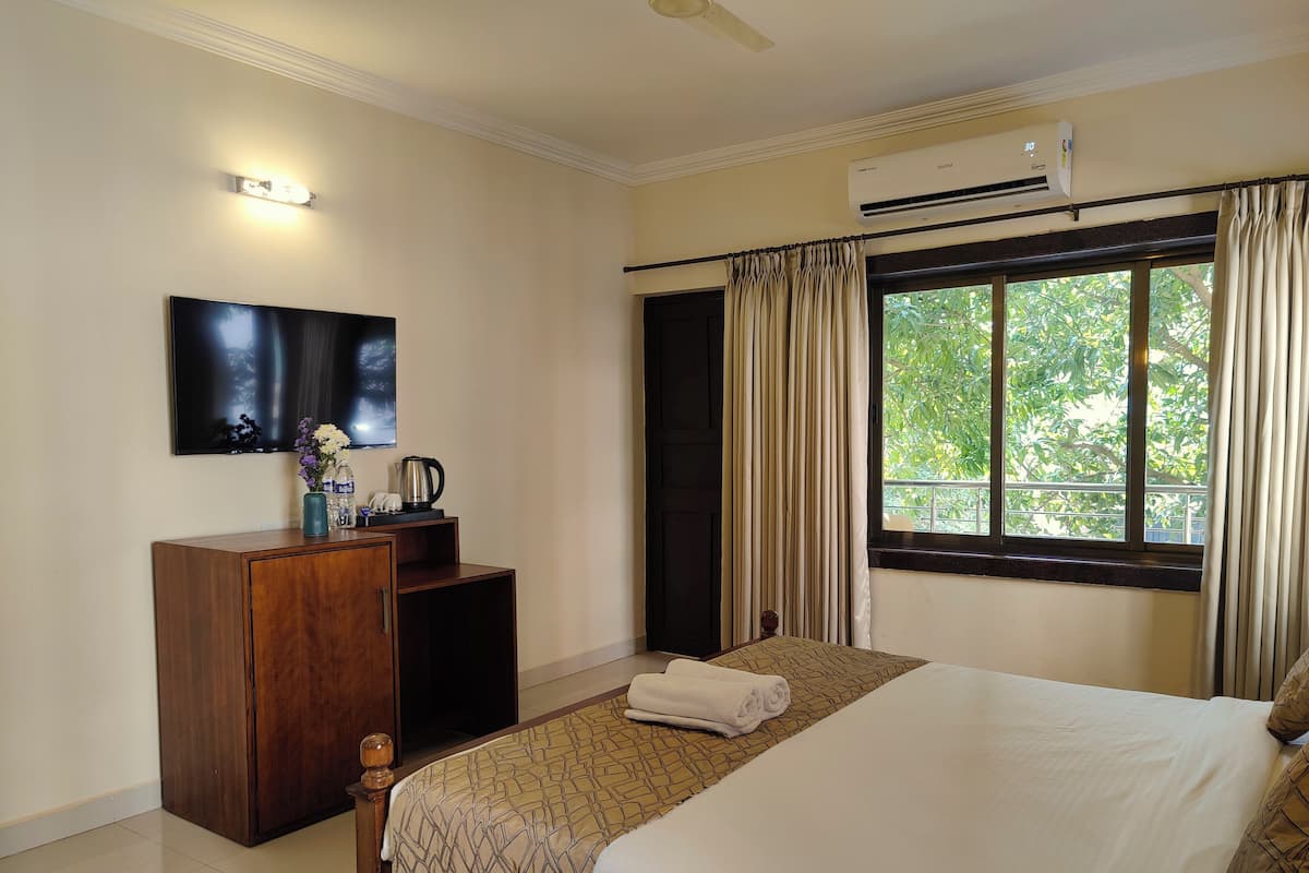 A hotel room with a bed, a TV mounted on the wall, a wooden dresser, a window with curtains, and an air conditioning unit.