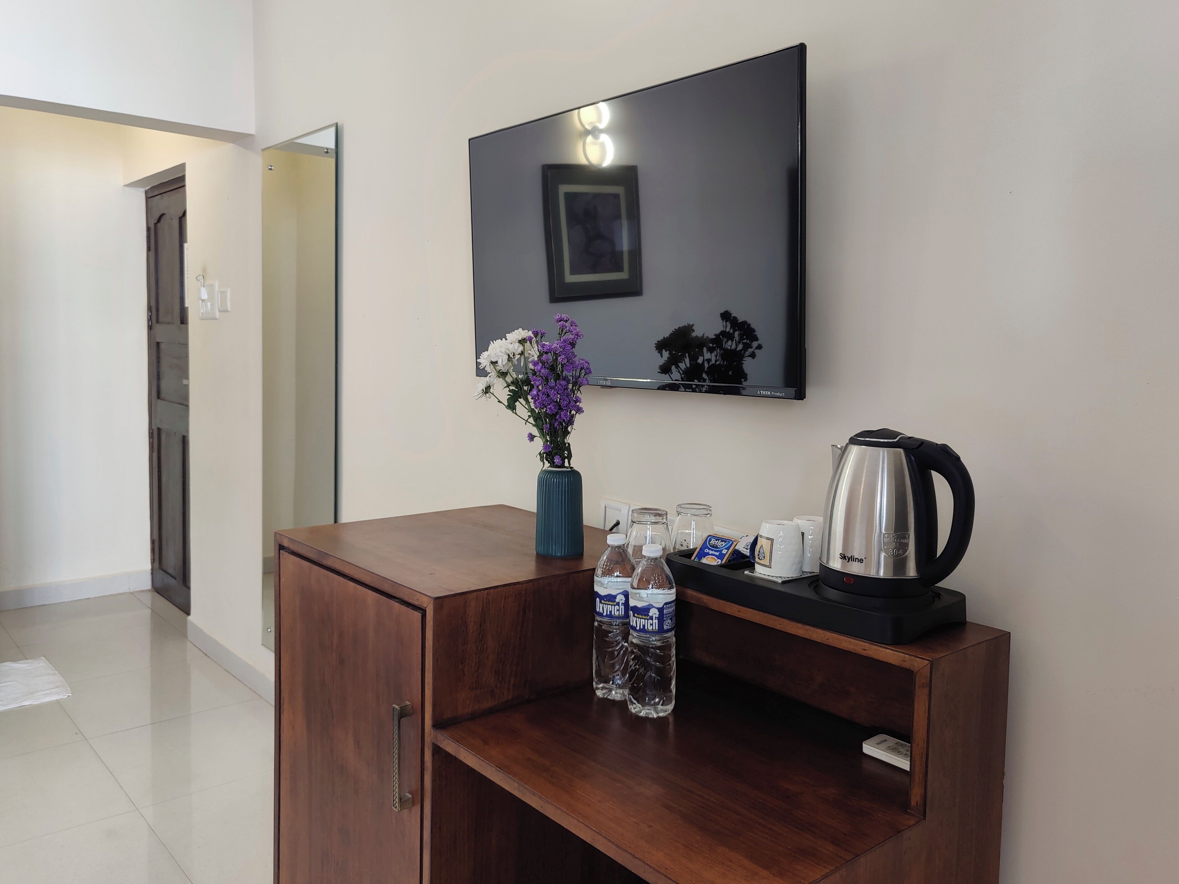 Habitación de hotel con un escritorio de madera, un florero con flores, una tetera y un televisor en la pared.