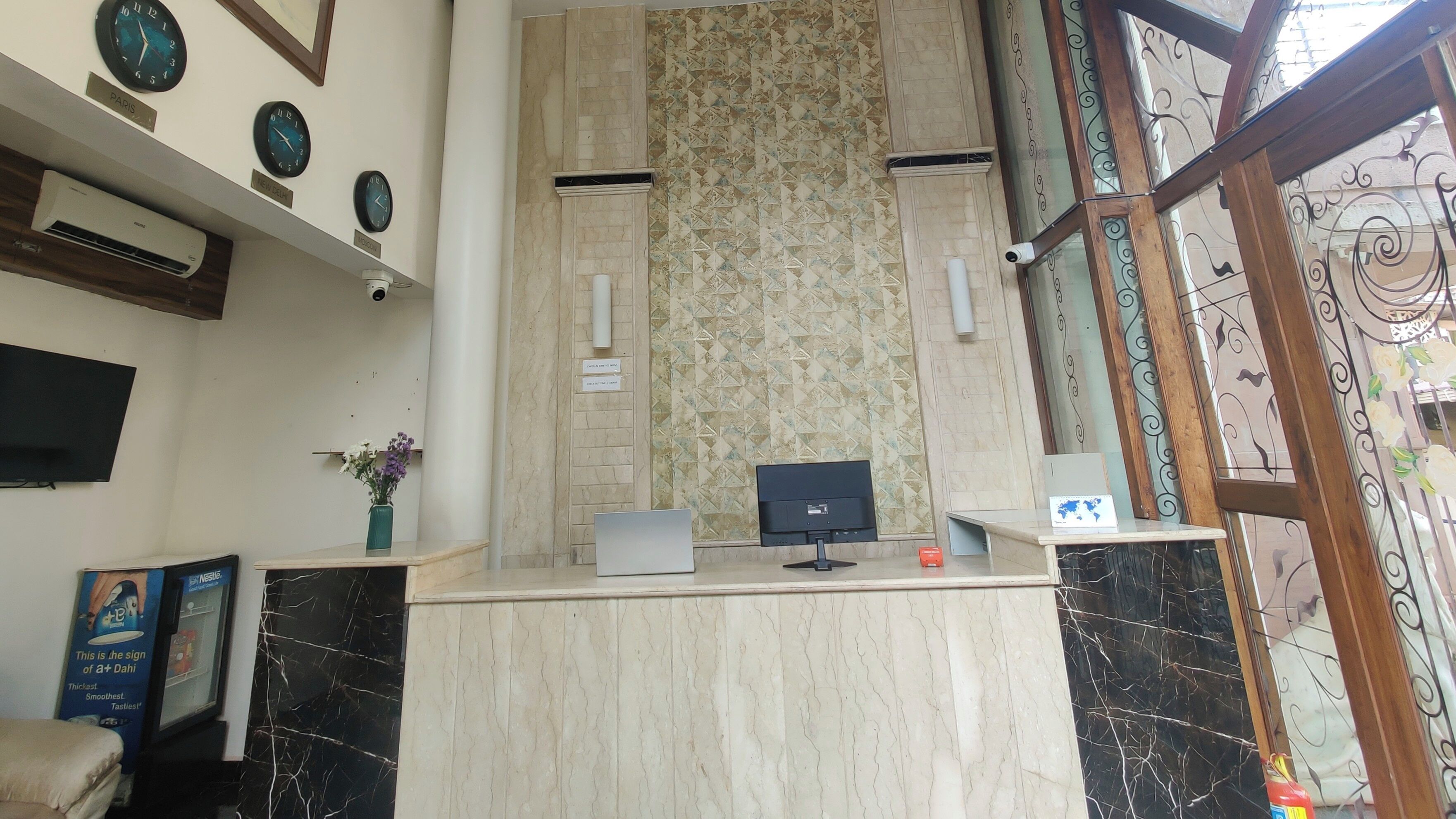 Un mostrador de recepción con un monitor de computadora, una lámpara de escritorio pequeña y un mapa del mundo. En la pared hay relojes con las etiquetas 'París' y 'Nueva Delhi'.