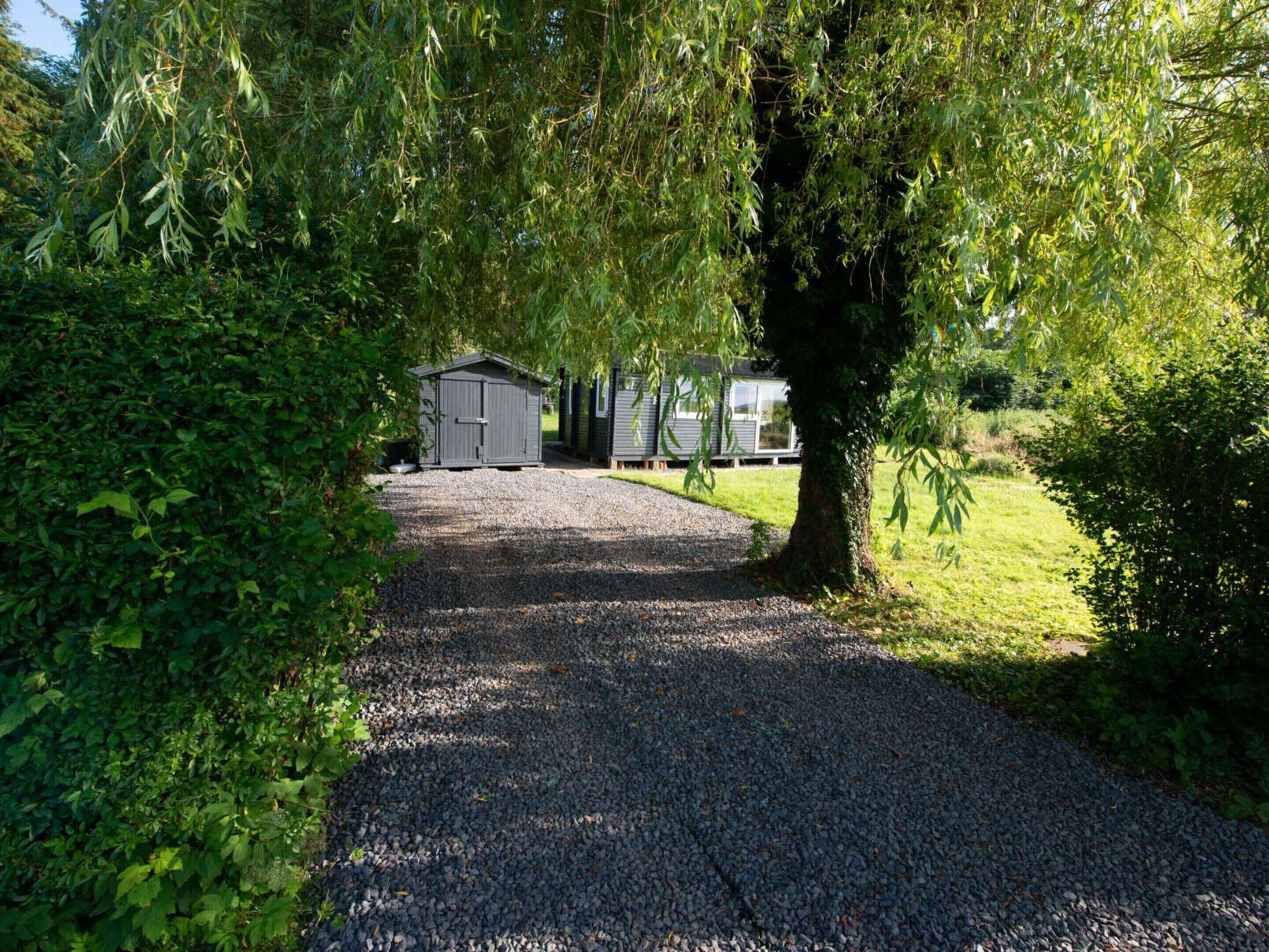 Ein Schotterweg führt zu einer kleinen Werkstatt und einem Haus, umgeben von üppiger Vegetation.