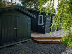 A wooden deck leading to a dark-colored shed with a door and window, surrounded by gravel and greenery. - 5 Star Holiday Home in Holbaek (Holbaek)