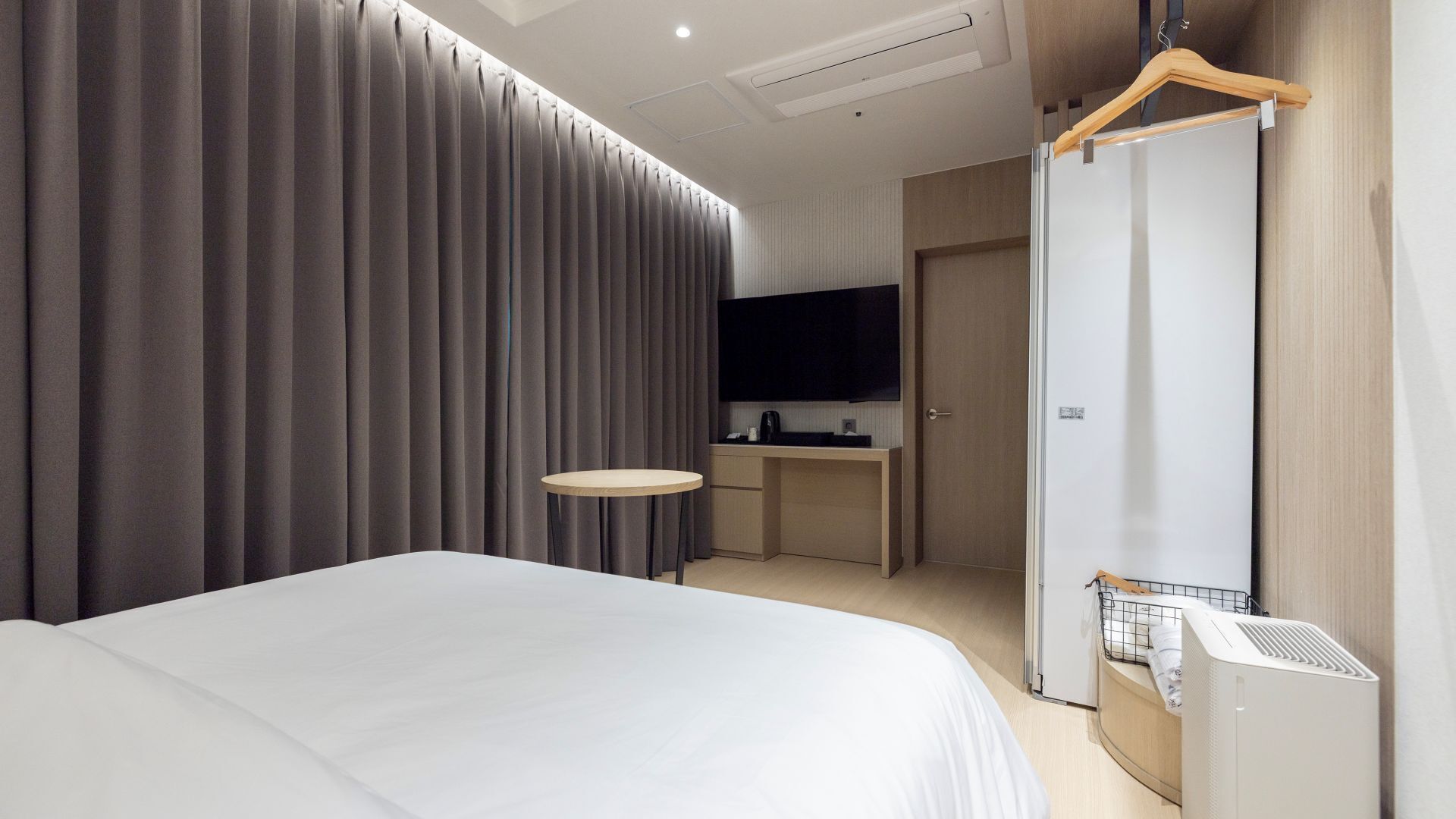 A modern hotel room with a large bed, a small round table, a flat-screen TV, a wardrobe, and a compact kitchenette.