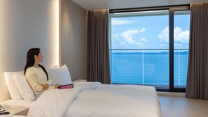 Una habitación de hotel con una cama grande, una mesita de noche con teléfono y control remoto, y vistas al mar a través de ventanales de gran altura.