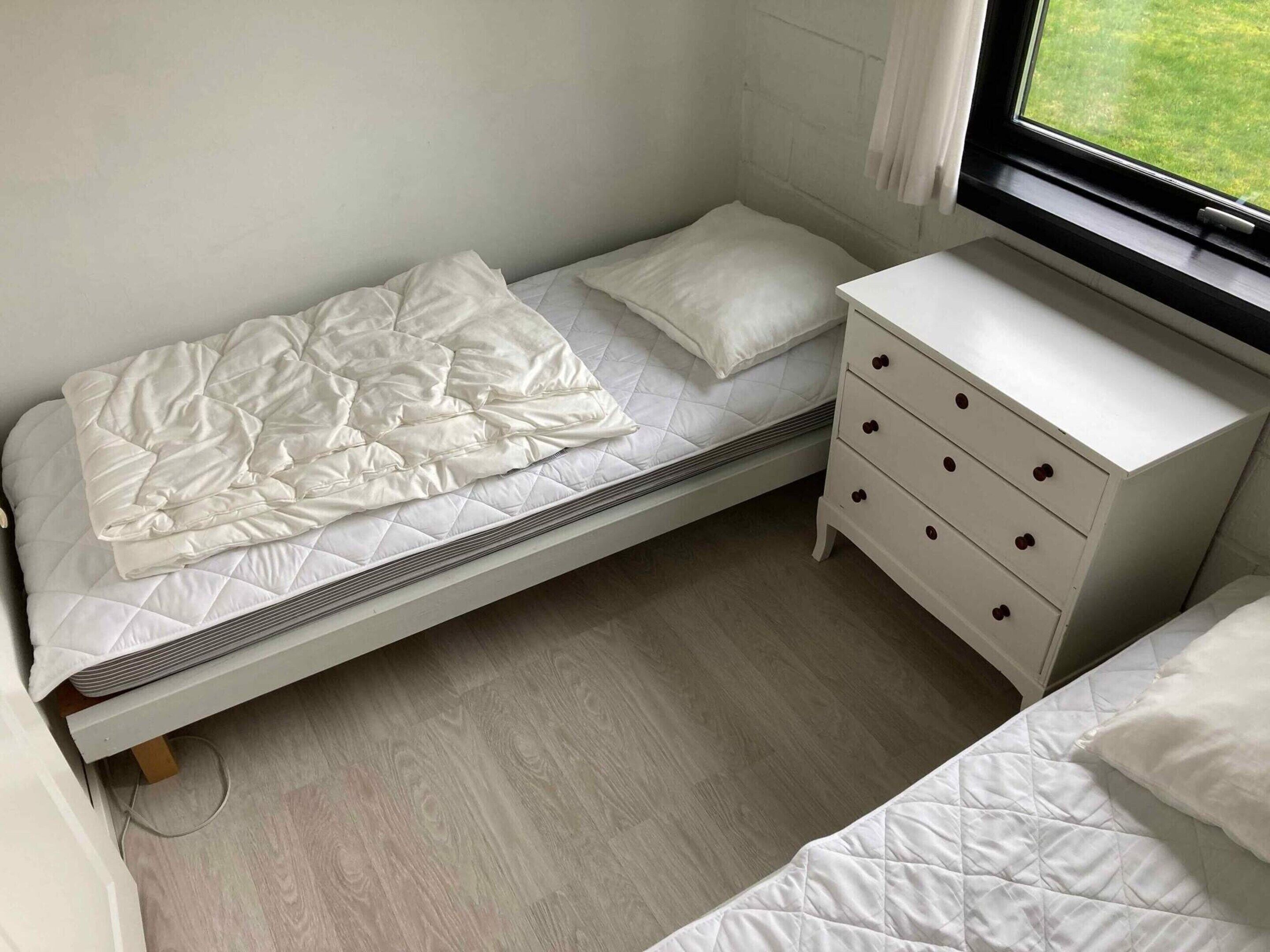 A small, single-bed room with a wooden floor, a white dresser with drawers, and a window with a view of a grassy area.