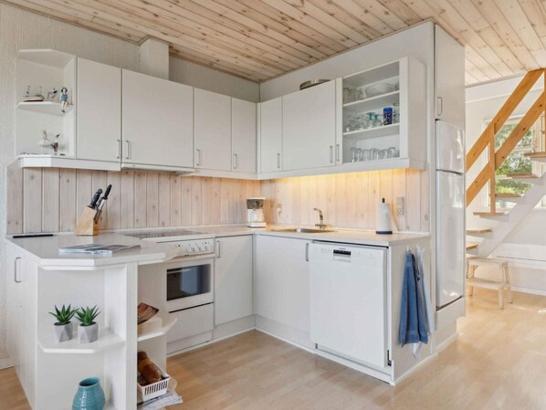 A modern kitchen with white cabinetry, a built-in oven, and a dishwasher. There is a wooden ceiling and a staircase leading to another level.