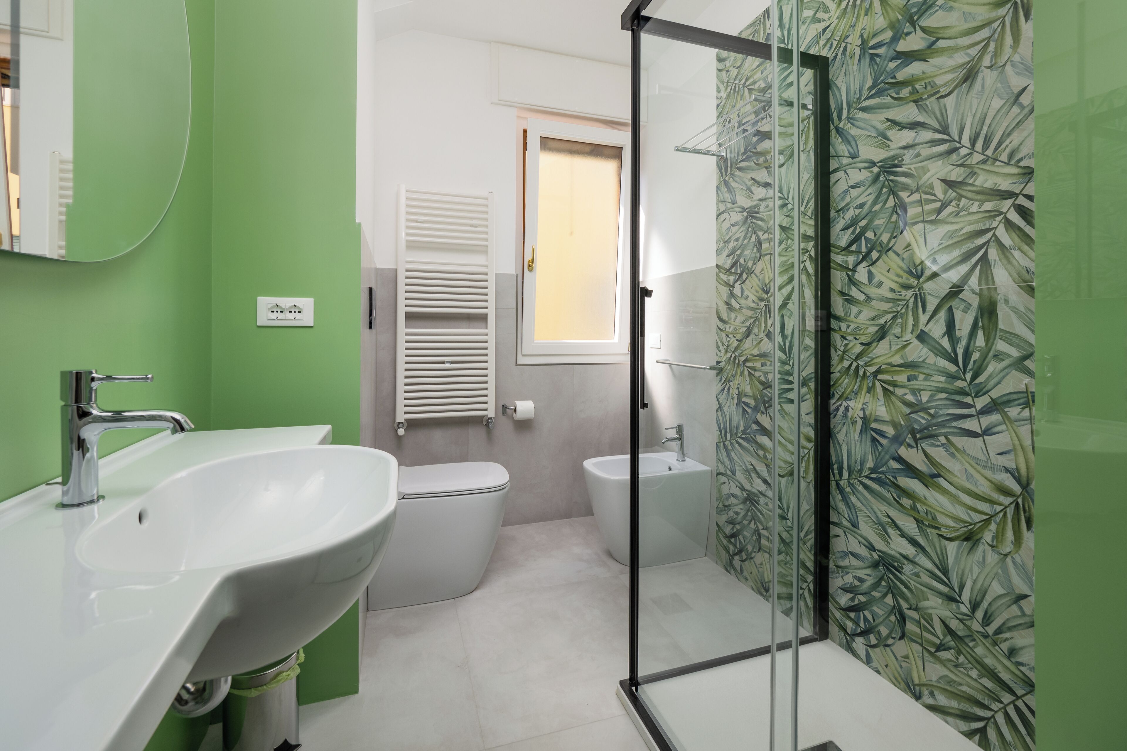 Un baño con lavamanos, inodoro y bidé blancos, cabina de ducha de vidrio y una ventana con persianas.