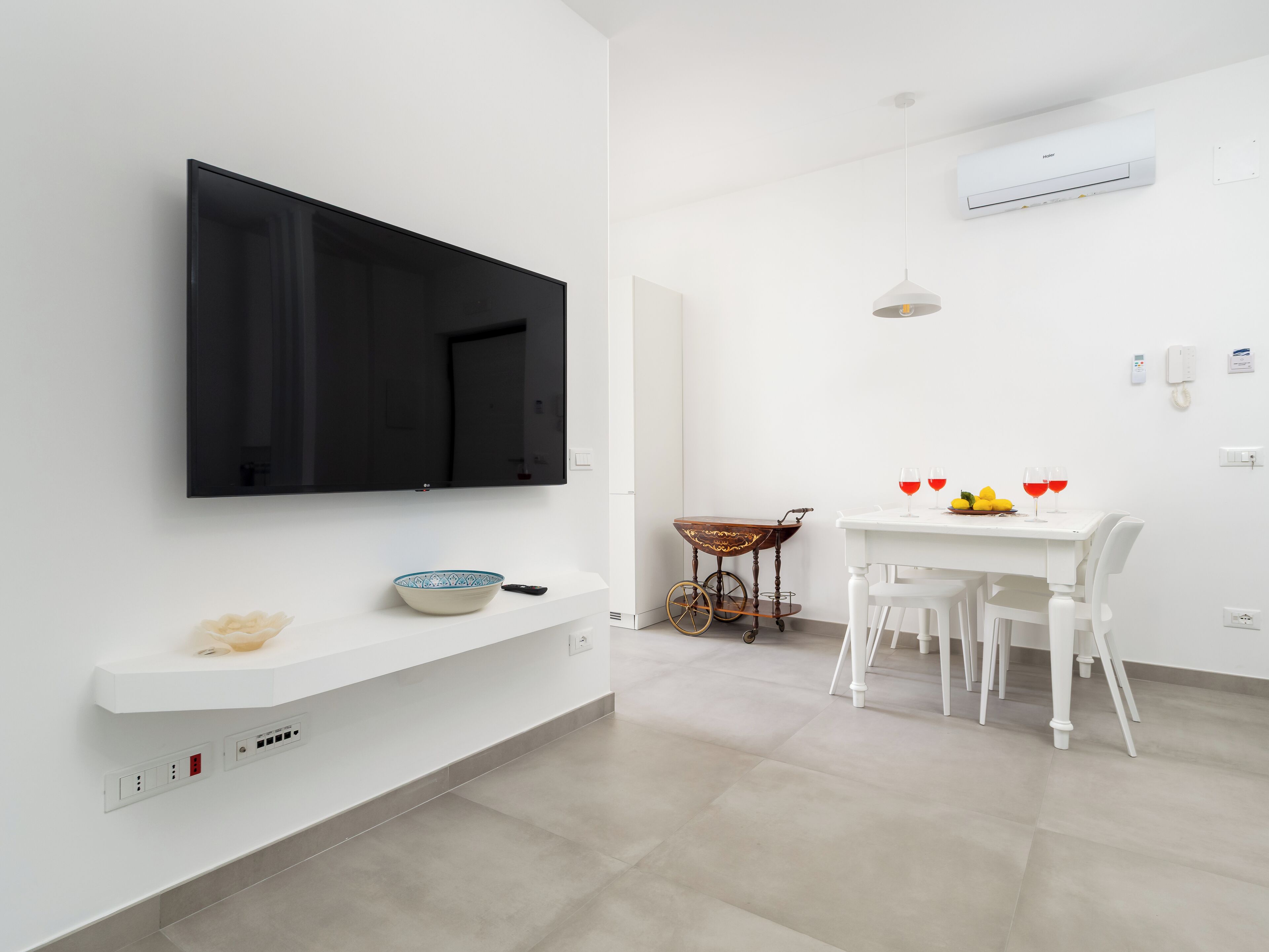 Een moderne woonkamer met een flatscreen televisie aan de muur, een witte salontafel met een schaal en afstandsbediening, een eethoek met een witte tafel en stoelen, en een klein karretje met wielen.