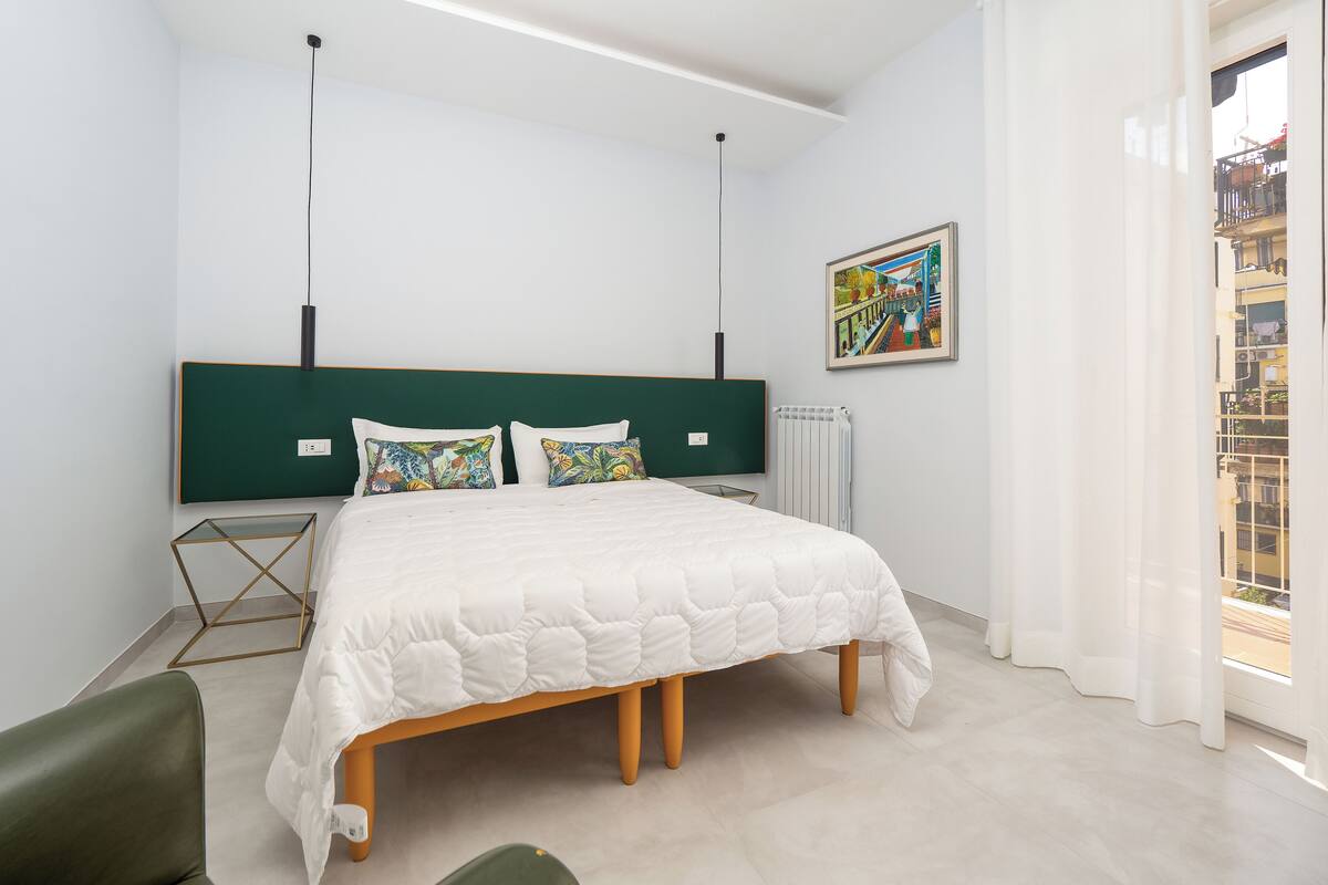 A modern bedroom with a large bed, a green headboard, a radiator, and a window with curtains.
