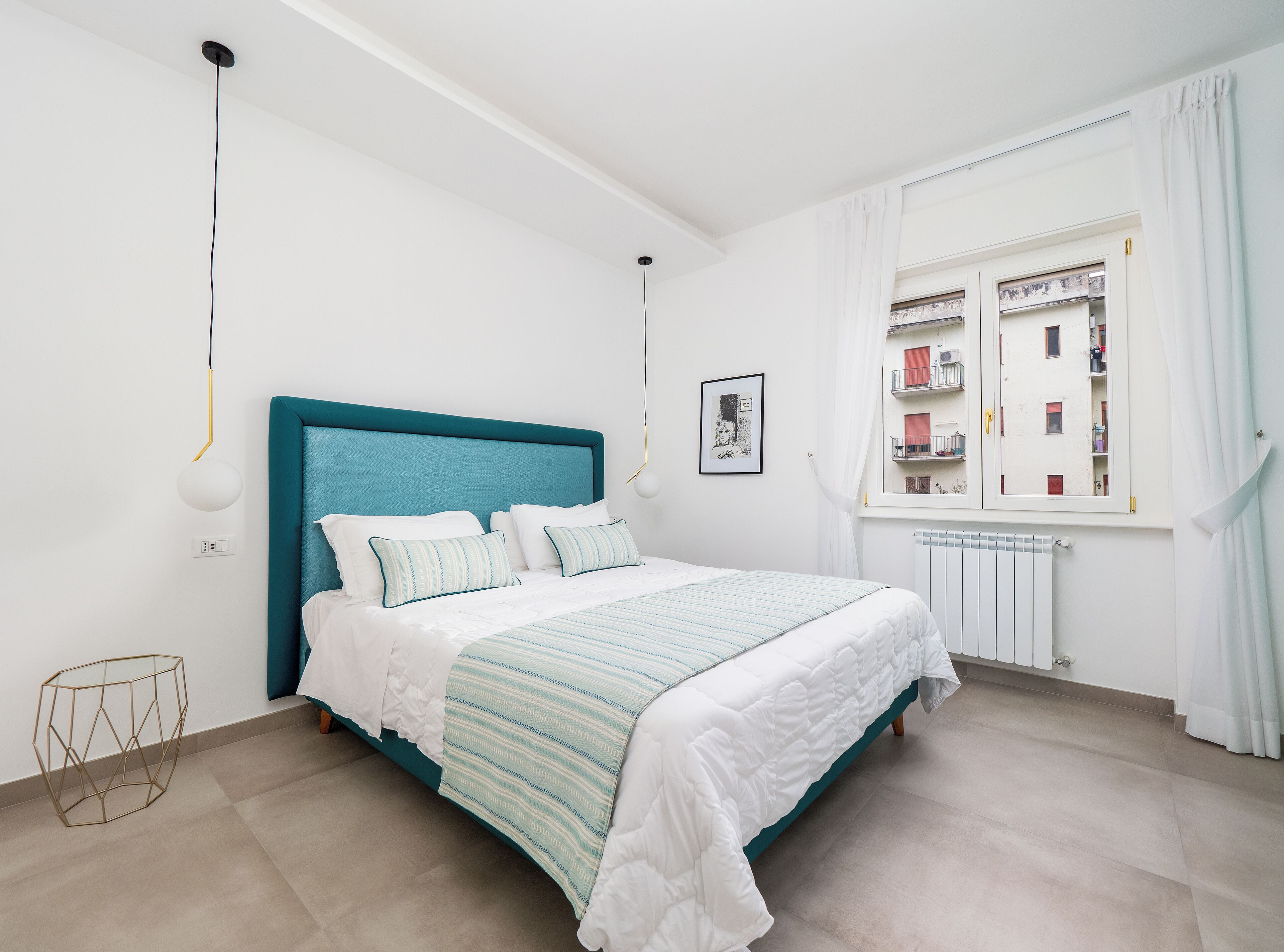 A bedroom with a teal headboard, white bedding, a geometric side table, and a window with a view of buildings.
