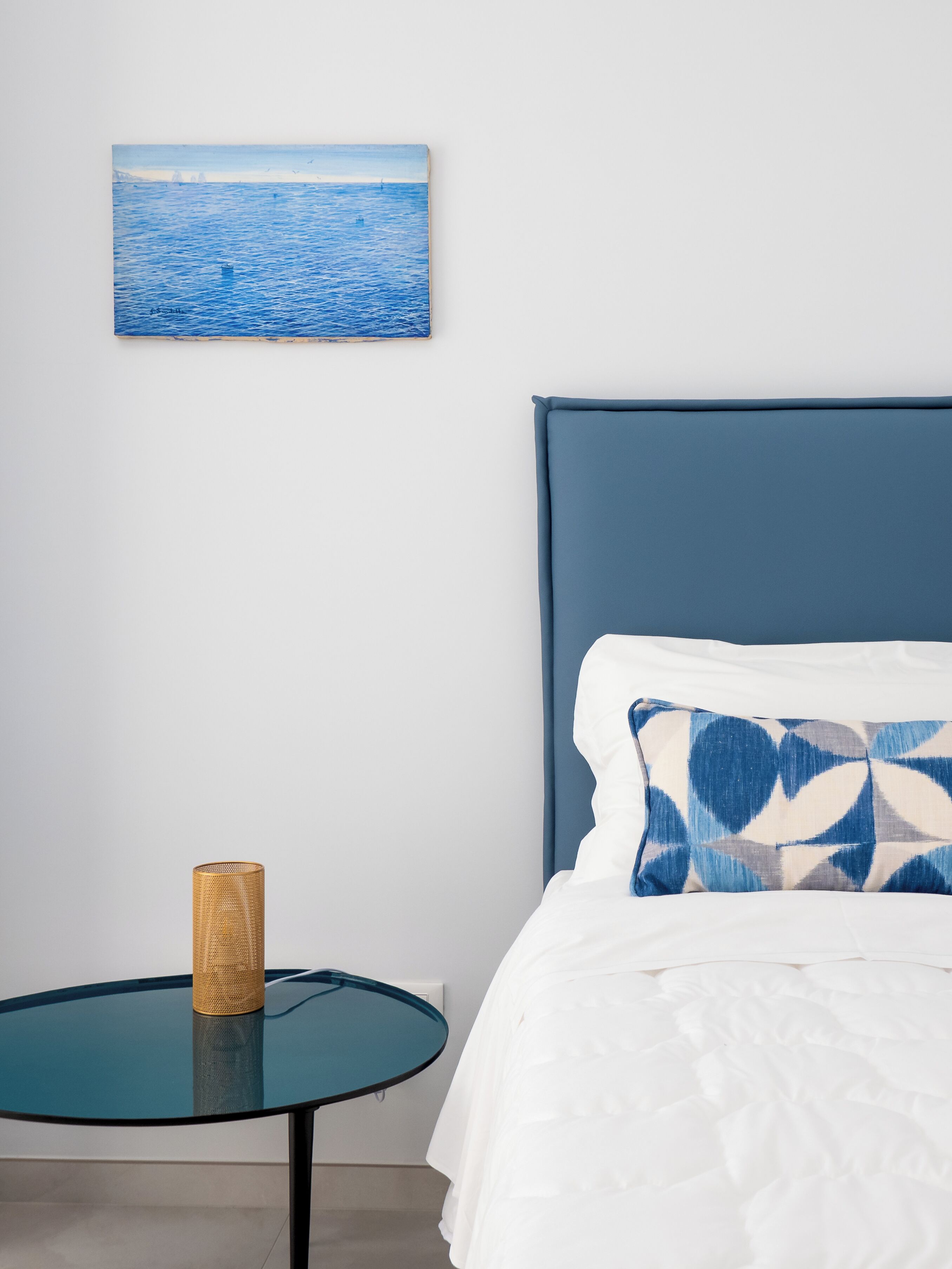 A bedroom with a bed, a round glass table with a woven basket, and a painting of a sea view on the wall.