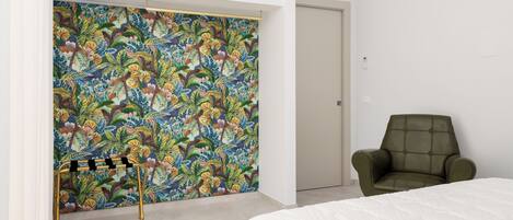 Un dormitorio con cama, un sillón verde, un muro con estampado floral y un armario blanco.