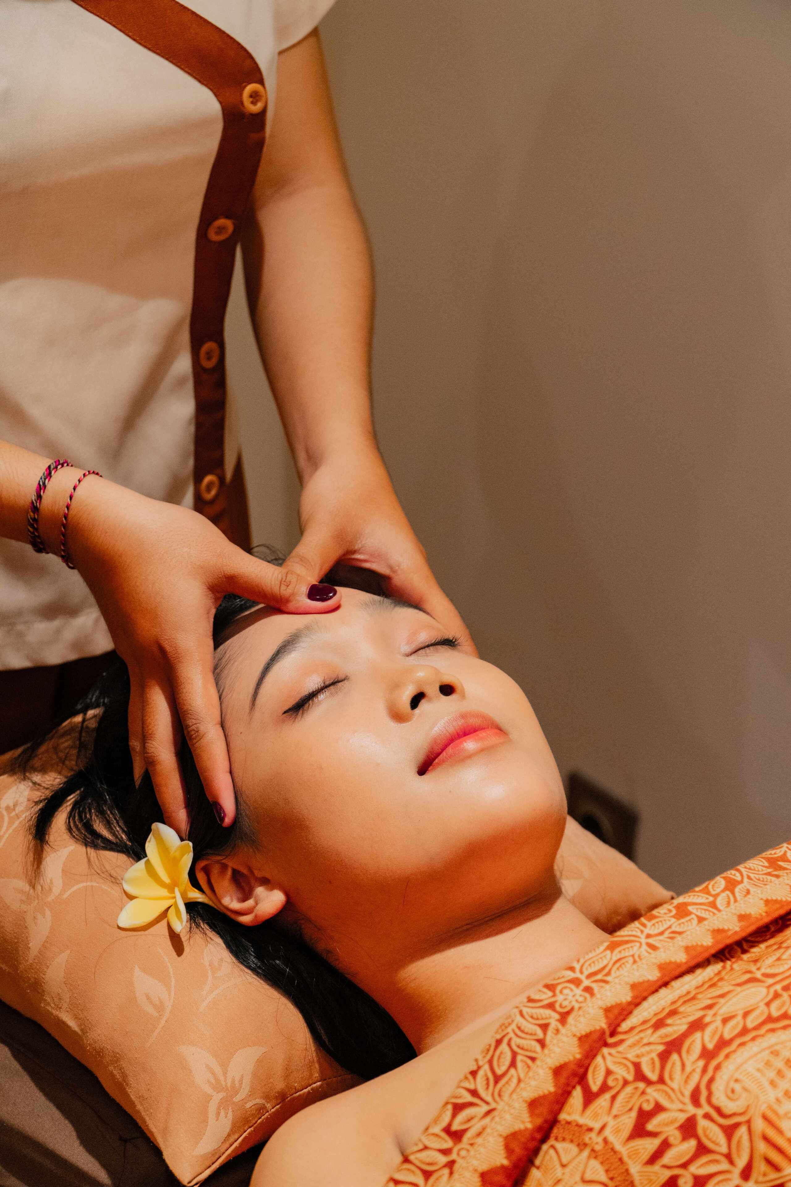 Eine Person liegt entspannt mit einer Blume im Haar und erhält eine Kopfmassage.
