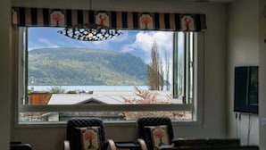 Een kamer met een groot raam met uitzicht op een meer en bergen, twee gestoffeerde fauteuils met bloemenkussens, en een hanglamp.