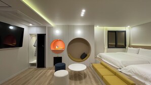 Quarto de hotel moderno com uma cama, um espaço de estar circular, uma mesinha e uma televisão.