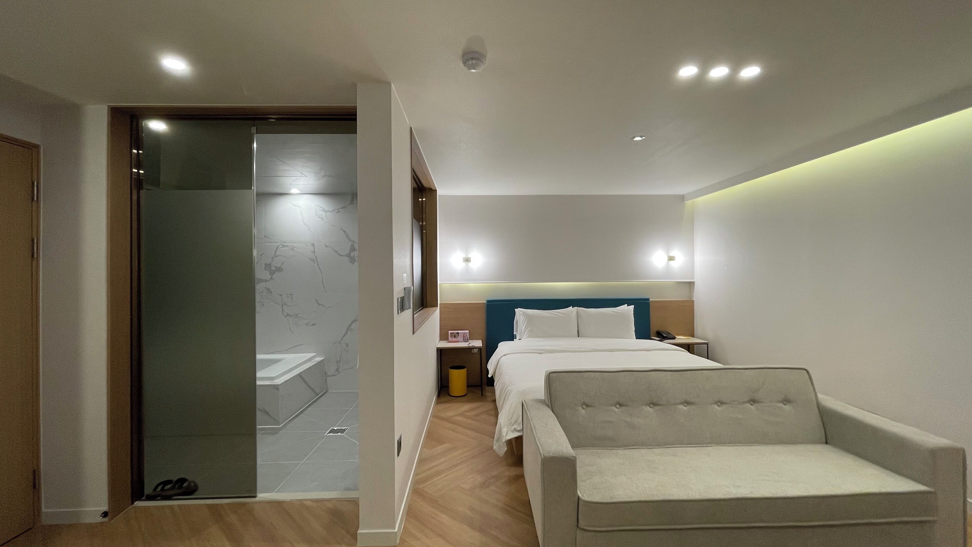 Habitación de hotel moderna con cama, una silla, una mesita de noche y un baño con puerta de vidrio.