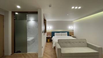 Habitación de hotel moderna con cama, una silla, una mesita de noche y un baño con puerta de vidrio.