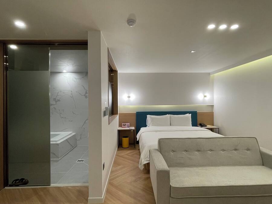 Habitación de hotel moderna con cama, una silla, una mesita de noche y un baño con puerta de vidrio.