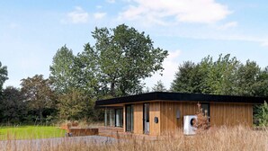 A modern, single-story cabin with a wooden exterior and large windows, surrounded by tall grass and trees. - 5 Star Holiday Home in Aakirkeby (Akirkeby)