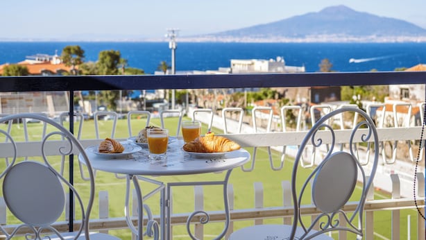 A balcony with a table set for breakfast, overlooking a coastal view with a mountain in the distance. - Casa Cafarelli (Sorrento)