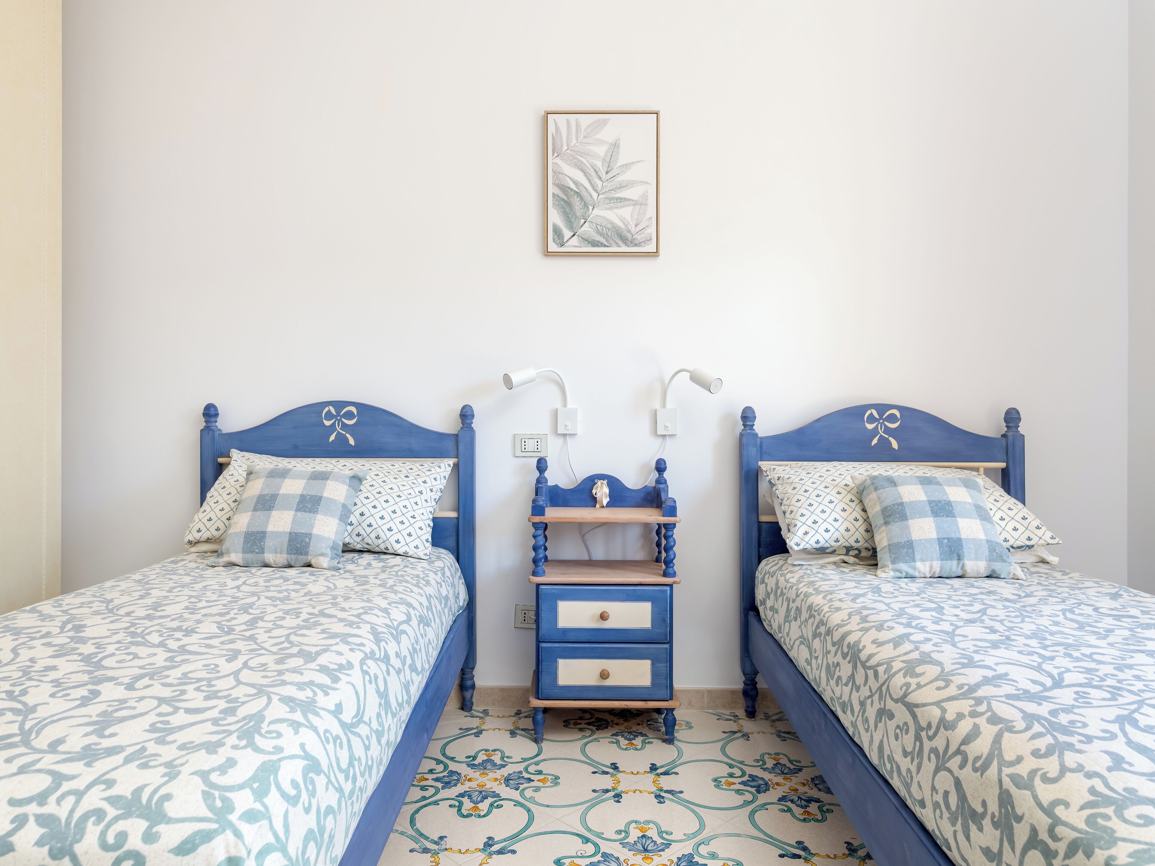 A bedroom with two beds, each with a blue headboard and matching pillowcases. There is a small wooden nightstand with drawers between the beds.