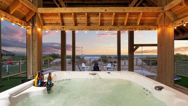 Un jacuzzi con una pérgola de madera encima, rodeado de sillas y vistas a la playa al atardecer.