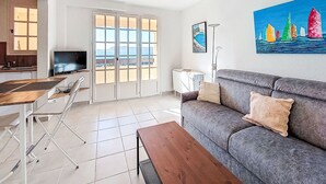 Una sala de estar moderna con un sofá gris, una mesa de centro de madera y un televisor en la esquina. Hay un balcón con ventanas amplias y un cuadro con barcos de vela en la pared.