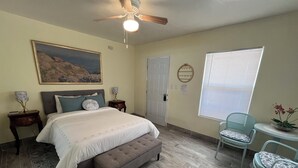 A bedroom with a bed, a bench, a chair, a ceiling fan, a window with blinds, a painting on the wall, and a small table with a lamp.
