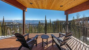 Um deck de madeira com cadeiras Adirondack e uma mesa pequena, com vista para uma paisagem montanhosa com picos nevados e florestas densas.