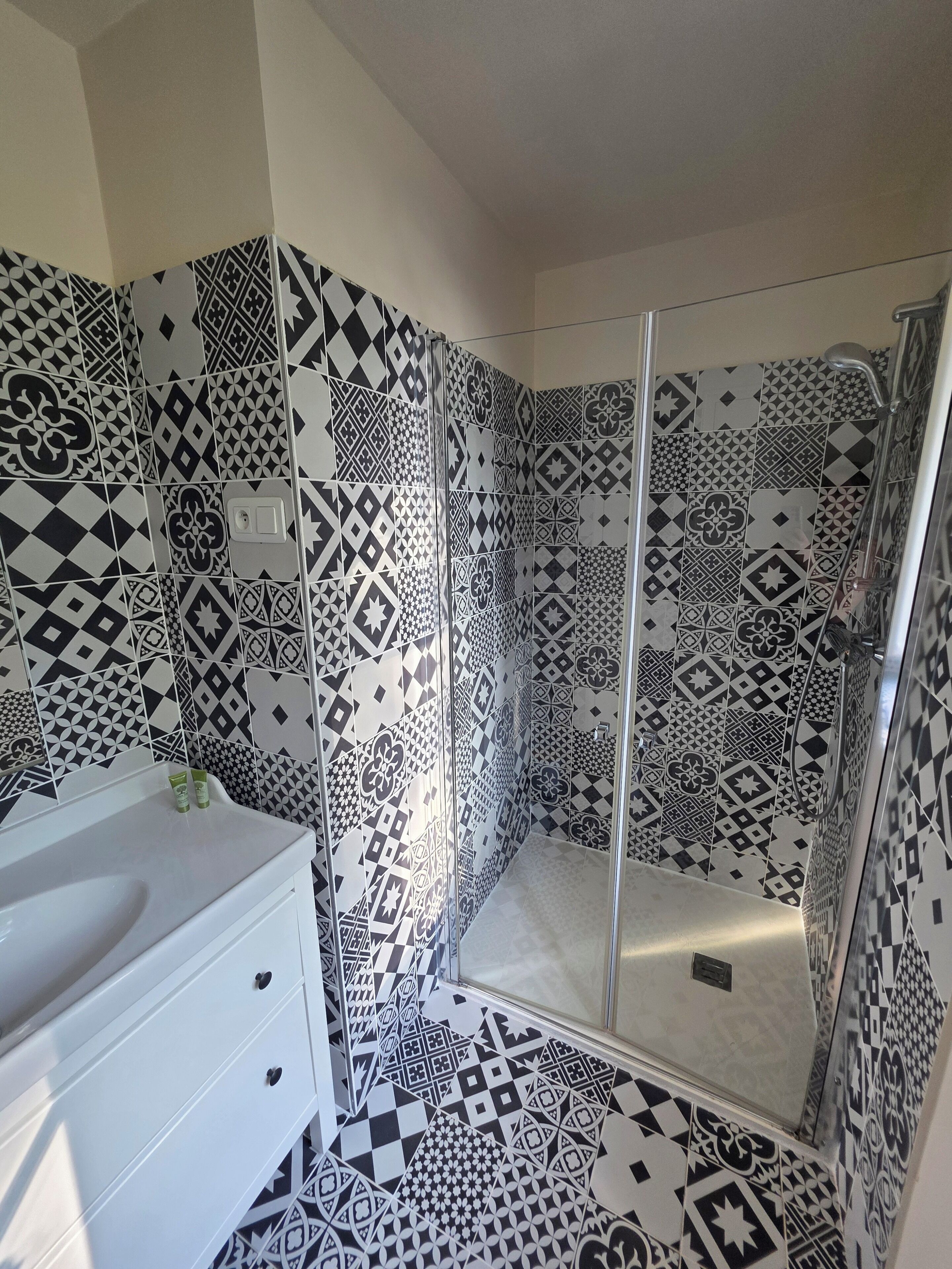 A bathroom with a shower area featuring black and white patterned tiles, a white sink with two green bottles, and a white cabinet with drawers.