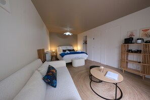 A modern hotel room with a sofa, a small round table, a bed with a blue blanket, a wooden shelf with kitchen items, and a ceiling fan. - Manoir de l'Échauguette  (Laignes)