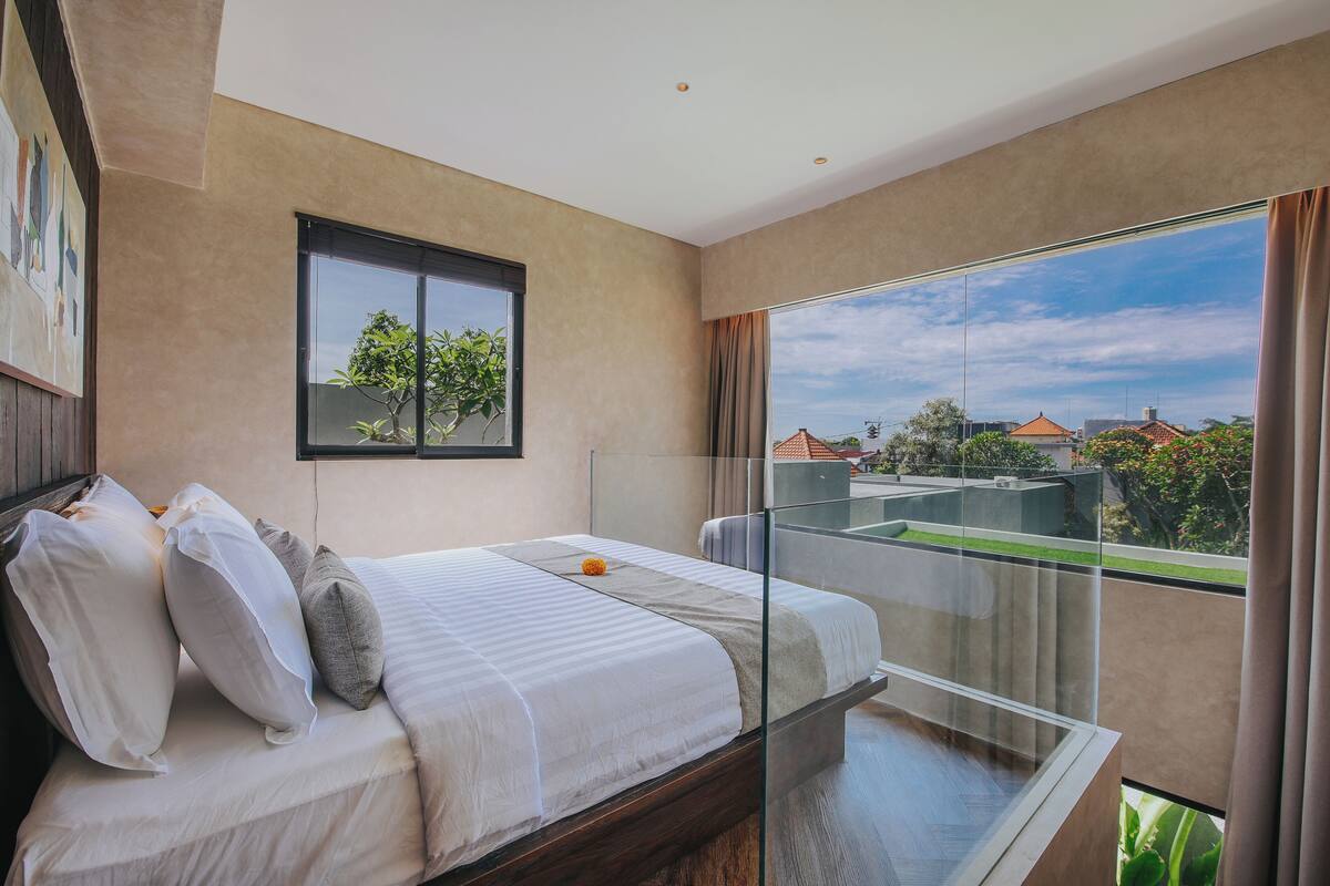 A modern hotel room with a large bed, a glass balcony, and a view of the cityscape.