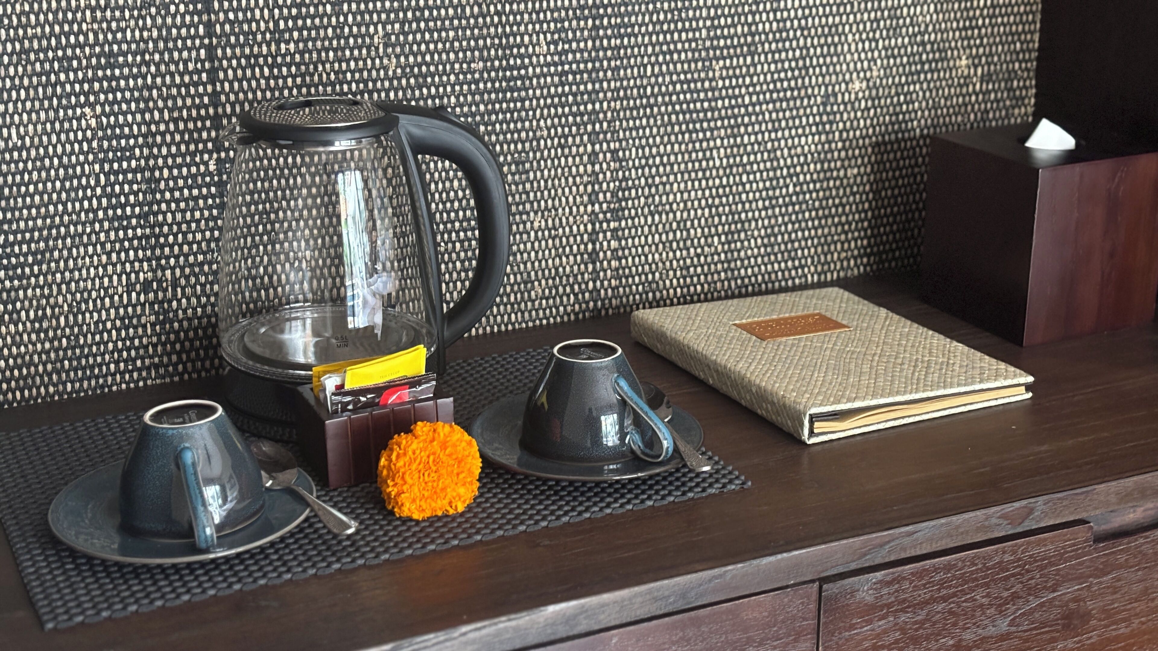 Una encimera de cocina con una cafetera, dos tazas, un libro y una flor naranja decorativa.