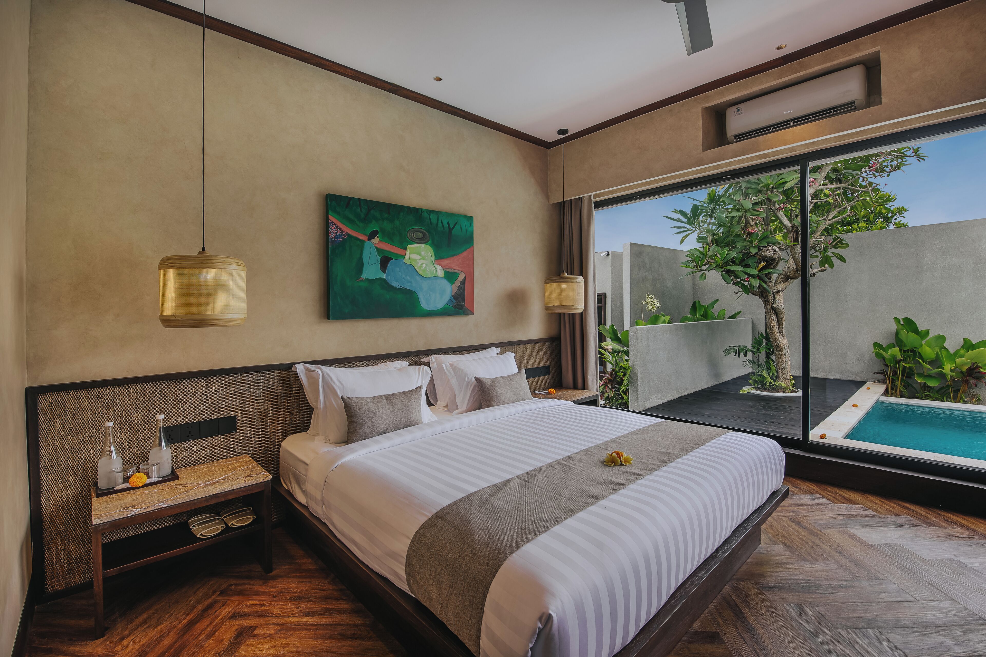 A modern hotel room with a large bed, a bedside table with a bottle and glasses, a painting on the wall, and a view of a swimming pool and greenery.