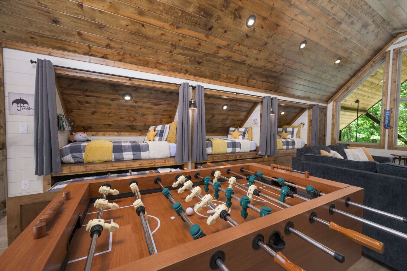 Une pièce avec un plafond en bois, deux lits avec une literie à carreaux, une table de baby-foot et un canapé.