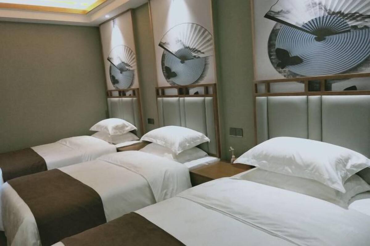 A hotel room with three single beds, each with a brown headboard and white bedding. There are wall-mounted fans and a large abstract art piece above the beds.